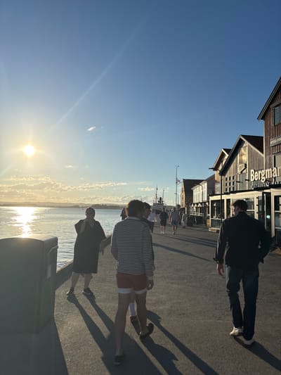 The image shows a sunny day on a waterfront promenade in Tønsberg, Norway. A number of people are walking along the promenade, which is lined with buildings. The sun is low in the sky, casting long shadows. In the background, there are boats docked in the water and the town of Tønsberg visible across the bay. One building prominently displays a sign for "Bergman Restaurant". The scene appears to be lively, with several individuals engaged in casual activity along the waterfront.