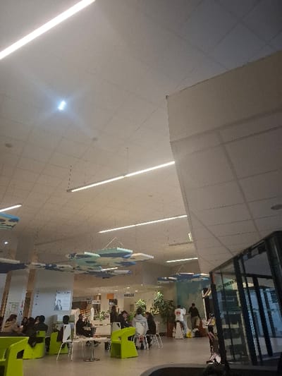 A brightly lit, modern indoor space features a high, white ceiling with parallel fluorescent lights. Below, several people are seated at tables with green chairs, appearing to be reading or studying. Decorative blue and white abstract shapes hang from the ceiling. A bookstore is visible in the background with people shopping. The scene is slightly rotated, presenting a tilted perspective.