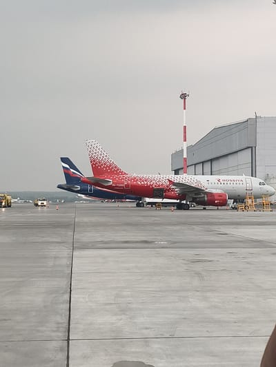 An Airbus A320-200 aircraft, registration RA-73203, operated by Rossiya Airlines, is parked on the tarmac at Emelyanovo Airport (KJA) in Emelyanovo, Russia. The aircraft features a distinctive red and white livery with a pattern of geometric shapes on its tail and rear fuselage. The Rossiya logo is visible on the forward fuselage. The aircraft is positioned in front of a large hangar-like structure. Several yellow service stairs are attached to the aircraft's fuselage, indicating maintenance or servicing activities. In the background, a red and white striped control tower or antenna is visible. Other airport vehicles, including a yellow tow truck and a white utility vehicle, are present on the tarmac. The scene is captured on a cloudy day, with a gray sky and wet tarmac, suggesting recent rain. The foreground consists of a concrete apron with visible seams and tire marks.