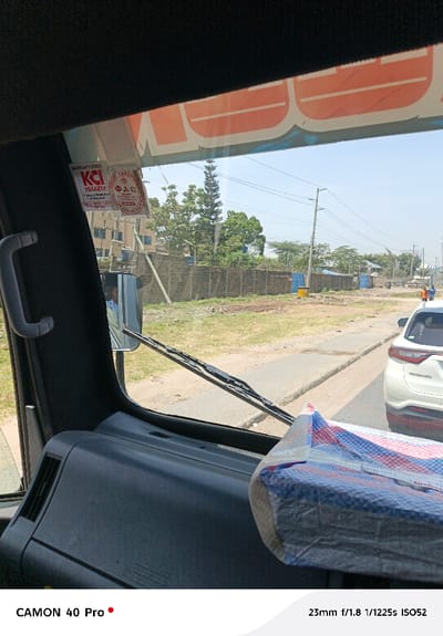The image shows a view from inside a vehicle, looking out through the windshield. A white car is visible on the road ahead. A grey wall runs alongside the road, and greenery is visible beyond it. A "WARRANTY STICK ISUZU" sticker is affixed to the windshield. A package wrapped in blue and white plastic sits on the dashboard. The camera information "CAMON 40 Pro 23mm f/1.8 1/1225s ISO52" is displayed at the bottom.