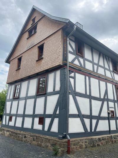 The image shows a section of a traditional German half-timbered house in Neustadt, Germany. The house has a dark gray timber frame with white infill panels. The roof is covered in small, overlapping tiles, a common roofing material in older European architecture. Several windows with dark frames are visible, some with multiple panes. There's also a gutter system running along the roofline, with a decorative element on one section featuring red and yellow painted ornamentation. The house is built against a rough stone wall on one side, and a cobblestone pathway is visible on the right edge of the image. The sky is overcast, suggesting a cloudy day. No people are visible in the frame. The architectural style and cobblestone suggest a historical setting. The time of day is likely daytime, given the visible details and light.