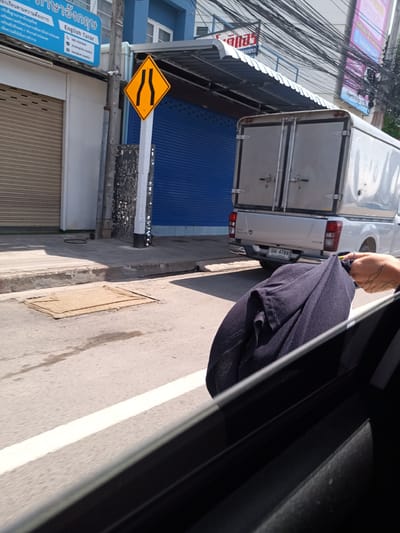 The photograph, captured from inside a vehicle in Hua Hin City Municipality, Thailand, presents a street scene. The view includes a yellow road sign indicating the narrowing of the road, adjacent to a building with a blue sign that reads "English Tutor." A pickup truck with a silver-colored cargo bed is visible on the right side of the frame, its license plate reading "บม 4769." The road is marked with white lines, and a sewer cover is visible on the asphalt. A person's hand and a dark blue garment are in the foreground, partially obscuring the view from inside the car. Overhead power lines are also visible.