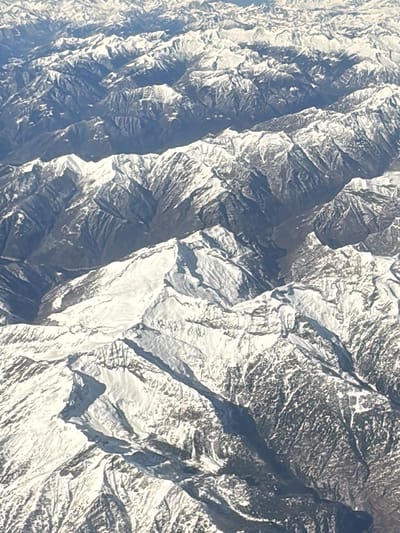 The image presents a stunning, high-altitude aerial view of a vast, snow-covered mountain range, likely captured from an airplane. The landscape is dominated by numerous peaks and ridges, all appearing rugged and imposing.

The mountains are extensively covered in white snow, particularly on their upper slopes and jagged summits. This snow glistens under bright daylight, suggesting clear weather conditions. The white snow starkly contrasts with the darker, exposed rock faces and the deep blue-grey shadows that fill the valleys and lower slopes. These shadows emphasize the dramatic contours and immense scale of the terrain, revealing deep ravines and steep inclines.

The light source, presumably the sun, appears to be positioned such that it illuminates one side of the mountain ranges, casting long, distinct shadows on the opposing sides and into the valleys below. This creates a strong sense of depth and three-dimensionality. The overall impression is one of a pristine, untouched wilderness.

The mountain ranges stretch across the entire frame, receding into the distant horizon where they eventually blend into a lighter, hazier blue, indicating the vastness of the scene. There are no visible signs of human activity, structures, roads, or significant vegetation, highlighting the remote and desolate nature of the high-altitude environment. The image conveys a sense of grandeur, tranquility, and the raw power of nature.