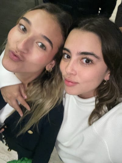 The specific location within La Purísima, Mexico, is not discernible from the provided image, which captures a close-up, eye-level selfie of two young adult females in an indistinct, dark indoor background.
The individual on the left has long, wavy light brown hair with blonde highlights and olive-toned skin. She wears multiple small gold hoop earrings in her visible left ear. Her lips are pursed in a 'kiss face,' and her light brown eyes are directed towards the camera. She is attired in a white high-neck garment layered under a dark, possibly navy or black, jacket featuring prominent gold buttons. Her right hand is positioned with fingers splayed, resting on the upper left arm or shoulder area of the individual on the right.
The individual on the right has long, wavy dark brown hair and fair skin. She wears a single small gold-colored drop earring in her visible right ear. Her expression is a subtle smile, and her dark brown eyes are directed towards the camera. She is wearing a white, textured, high-neck sweater.
The two individuals are positioned in close proximity, suggesting a shared photograph, likely a selfie. Lighting is uniform, casting no distinct shadows, consistent with an indoor environment.