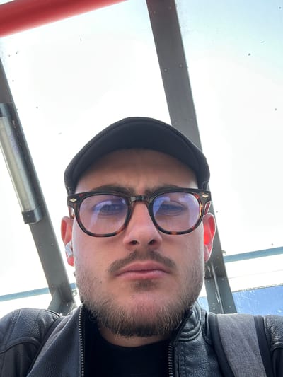 A close-up, rotated portrait captures an individual looking directly towards the camera from a low-angle perspective. The person has a light beard and mustache, and is wearing tortoiseshell-framed glasses featuring a small diamond-shaped embellishment on the side. A dark knit cap covers their head, and they are dressed in a dark leather jacket with grey fabric detailing on the sleeve. A white in-ear headphone is visible in their right ear.
The background is dominated by a bright, possibly overcast sky, suggesting daytime. Various architectural or structural elements frame the subject: a prominent silver-grey metal beam with visible rivets extends horizontally across the upper portion of the frame, accompanied by another narrower grey beam. A vibrant red vertical pipe is visible on the left side. Small specks, possibly dust or condensation, are scattered across what appears to be a transparent surface in the background. Reflections in the glasses show the bright sky. The setting, located in Geldern, Germany, appears to be an outdoor or semi-enclosed space, potentially a station or a structure with a large overhead opening. The person's expression is neutral. No visible text is present.