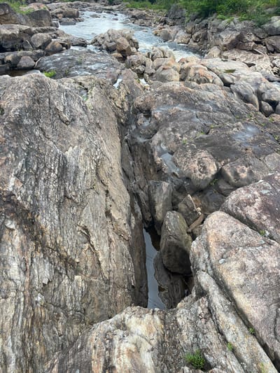 The image shows a rocky riverbed with flowing water and lush green vegetation on the banks in the background. The rocks are varied in size and texture, with some appearing wet and others dry. The water is clear and reflects the sky and surrounding rocks. The scene suggests a natural, untouched environment, possibly a park or wilderness area. The specific location in Village of Lyons Falls, United States of America is not identifiable from the image alone.