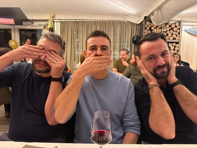 Three men pose in a "see no evil, speak no evil, hear no evil" manner. The man on the left covers his eyes. The man in the center covers his mouth. The man on the right covers his ears. All three are seated at a table with a partially filled wine glass in the foreground. Other diners are visible in the background. A balloon and stacked firewood are visible behind them.