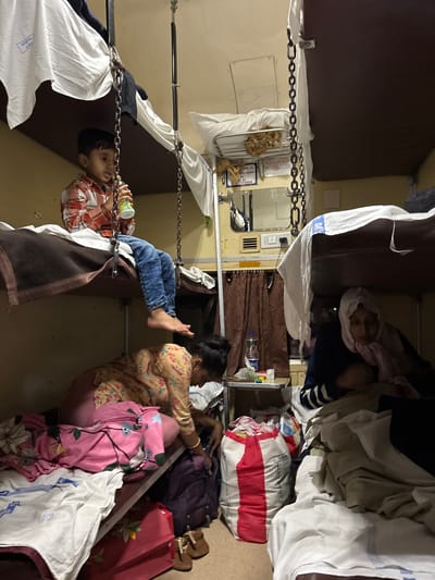The image depicts the interior of an Indian Railways train compartment. A young boy sits on a berth, drinking from a cup. A woman is seated on a lower berth next to luggage, including a red suitcase and bags covered with blankets and linens. An older woman, wearing a head covering, is on an upper berth amid more luggage. The scene includes chains suspending the upper berth, metal rails, and the characteristic beige interior of an Indian train. No information in the image corroborates the location being Ambernath, India.