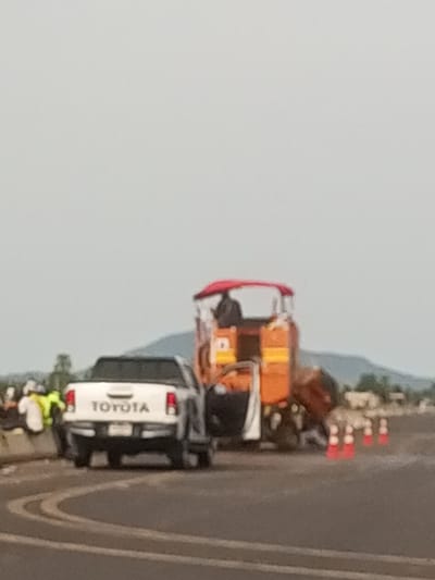 This media file captures a scene on a road in Baccure Kofare, Nigeria, with what appears to be road work or maintenance underway. The primary subjects are a white Toyota pickup truck parked on the left side of the frame and a large, orange construction vehicle on the right. The Toyota truck has its tailgate down, and a person in a yellow vest and white shirt is visible near it. The construction vehicle is orange with a red canopy and some yellow accents. It appears to be a type of paving or asphalt machine. Several orange traffic cones are lined up in front of the construction vehicle, indicating a work zone. In the background, there are some trees and hills under a hazy, overcast sky, suggesting it might be daytime with cloudy weather. The overall scene appears to be a typical road construction or repair site.