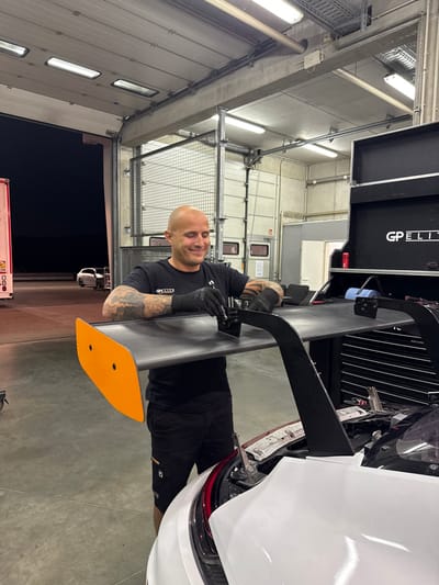 The image displays an automotive workshop or race team garage located in Hohenstein-Ernstthal, Germany, likely adjacent to the Sachsenring motorsport complex. The scene is artificially lit by overhead fluorescent fixtures, with a large garage door open to a dark, unlit exterior, indicating nighttime conditions.
A single male subject, bald and with extensive arm tattoos, is centered in the foreground. He wears a black short-sleeved t-shirt bearing the 'GP ELITE' logo and black work gloves. The individual is smiling and is actively engaged in adjusting or attaching a large, black, multi-element rear wing to the back of a white high-performance or race car. A bright orange endplate is visible on the left side of the rear wing. He is utilizing a small hand tool, possibly a wrench or hex key, on the wing's mounting hardware.
To the right of the subject, a black, multi-drawer tool cabinet or chest is present, featuring the 'GP ELITE' brand name prominently displayed in white text on its top section. The workshop floor is grey concrete. In the background, visible through the open garage door, a section of a white semi-trailer is present on the far left, and two white passenger vehicles are parked on a dark, paved surface under minimal ambient light in the distance.