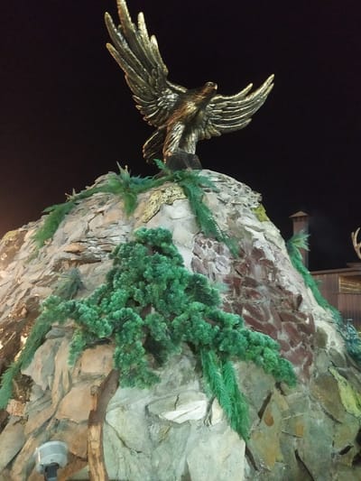 The image shows a nighttime exterior scene featuring a large, metallic eagle sculpture with outstretched wings perched atop a man-made rock formation. The sculpture is golden in color and detailed, appearing to be made of bronze or a similar material. The rock formation is textured and appears to be constructed from various stones and possibly brickwork, with artificial greenery resembling ferns and pine branches cascading down its sides. In the background, a dark sky is visible, along with hints of wooden structures and a chimney. A security camera is mounted on the lower left portion of the rock formation. The location is identified as Sosnovka, Kyrgyzstan.