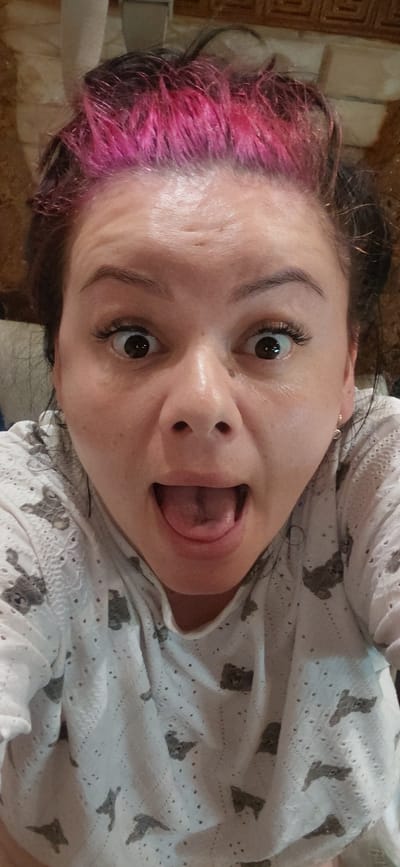 The media file is a portrait of a young woman with her face close to the camera, oriented sideways. She has dark hair with vibrant pink tips. Her mouth is open, and her tongue is sticking out. Her eyes are wide open, and she appears to be making a playful or surprised expression. She is wearing a white top with a subtle pattern of small, dark gray teddy bears. The background is blurry, but appears to be an indoor setting, possibly a home, with what looks like tiled wall. The lighting suggests it could be daytime.