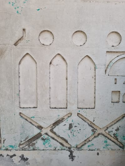 The image displays a weathered, light-colored wall surface, specifically located in Pedda Kammavaripalli, India. The wall is constructed of a material resembling stucco or plaster, coated in an off-white or light beige paint. Significant deterioration is evident, characterized by extensive paint peeling, revealing patches of faded teal or greenish-blue undertone and a darker greyish substrate. Fine cracks traverse the surface, and dark, irregular stains, possibly dirt or mold, are visible, particularly along the bottom edge and within recessed areas.
Geometric patterns are carved or indented into the wall's surface. From left to right, the top section features a distinctive hooked shape, followed by three circular indentations of uniform size, and a partial, larger circular or curved shape on the far right. Below these, a row presents three prominent, vertically oriented, arched rectangular indentations, each tapering to a point at the top, reminiscent of Gothic or Islamic archways. To the right of the third archway, a smaller, curved indentation partially frames a rectangular recess. The bottom section of the wall features large 'X' shapes, formed by intersecting diagonal grooves. Two complete 'X' forms are visible, with a partial third 'X' on the far left. All carved elements exhibit shadowed edges, indicating their recessed nature and depth. No people, animate subjects, or explicit actions are present within the frame. The environment is implied as an exterior or interior space with significant exposure to elements, contributing to the wall's aged condition.