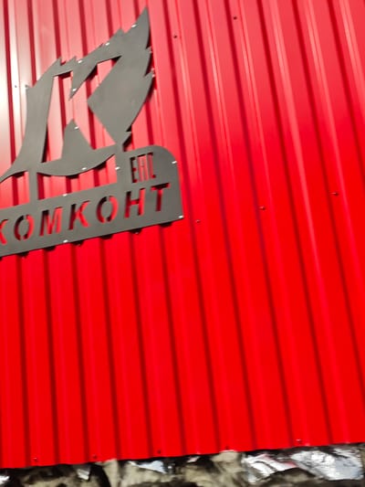 The image shows a close-up of a red corrugated metal wall with a black metal logo and text. The logo is abstract and sharp, with elements resembling flames or a stylized letter "K". Below the logo, the word "KOMKOHT" is visible, cut out from a black metal plate, with the letters "EAC" also cut out above it. The metal sheeting has vertical ridges. The lighting suggests it is daytime, with some sunlight casting shadows and highlights on the metal surface. The background is out of focus, but shows more of the red corrugated wall and a reflective, possibly insulated, material at the bottom of the frame. There are no people visible in the image. The setting appears to be an exterior wall of a building, possibly a commercial or industrial one, given the robust material and signage. The location context provided is Gubithsky Selsovet, Belarus, which gives a geographical reference for the scene.