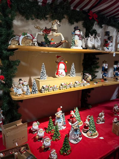A display of Christmas ornaments and decorations. A wooden shelf holds various figures, including snowmen, angels, and miniature trees, accented with greenery. Nutcrackers, sheep ornaments, and a carousel horse figurine also adorn the display. On a red surface nearby, Santa figures, decorative trees, and other ornaments are arranged, along with boxes.