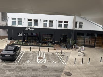 The image shows the exterior of "Le Burger" restaurant, located in Amstetten, Austria, indicated by the license plate on a black Volkswagen Tiguan parked in a handicapped parking space in front of the restaurant. The restaurant has a black facade with a white "Le Burger" sign. Above the restaurant, the windows of the building display various stickers for outdoor activities: "OUTDOOR", "SKI", "RUNNING", "BIKE". Outside seating is arranged under black umbrellas. A walkway with bollards leads to the restaurant entrance. Additional seating is visible, including chairs and benches. The ground is paved with grey bricks and several manhole covers. The overall scene suggests an urban setting with a mix of retail and dining.