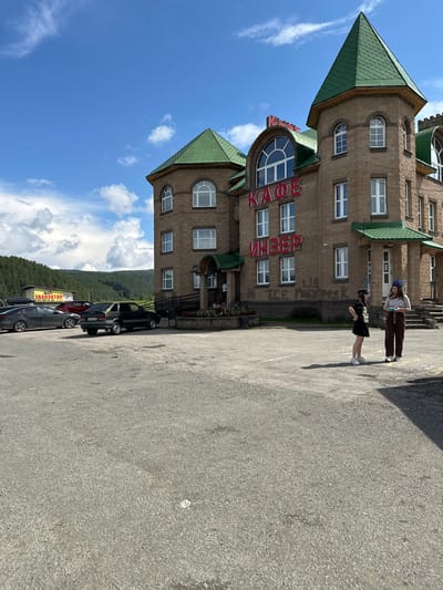 The image shows a large, multi-story building with a prominent green roof and towers, constructed from light-colored stone blocks. Red lettering on the facade reads "КАФЕ" (Cafe) and "ИНЗЕР" (Inzer), indicating it is a cafe named "Inzer." The building has numerous windows, many of which are arched. In front of the building is a large asphalt parking lot.

Two young women are standing in the parking lot, facing each other. One is wearing a black t-shirt, black skirt, black baseball cap, and white sneakers. The other is wearing a light-colored long-sleeved shirt, brown pants, and white sneakers.

Several cars are parked in the lot, including a black sedan and a dark gray sedan. In the background, a yellow banner with Cyrillic text that appears to be an advertisement or sign is visible. Beyond the parking lot, a forested hillside rises under a blue sky with scattered white clouds. The overall scene suggests a roadside cafe or establishment in a semi-rural or suburban setting. The location is confirmed by the signage as Inzer, Russia.