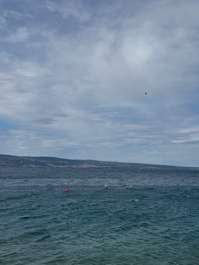 The image shows a view of the Adriatic Sea from the coast of Dugi Rat, Croatia. In the distance, a line of hills can be seen. The sky is mostly cloudy with a few patches of blue. A single bird is flying in the sky. The sea is choppy and has a dark blue color. Two red buoys are visible in the water. The environment is calm and serene. The coordinates of the location are 43.446 latitude and 16.634 longitude.