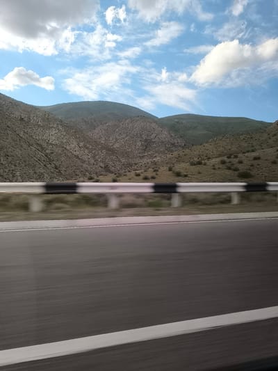 The image presents a dynamic view from a moving vehicle traversing a road through the mountainous landscape of Tigranashen, Armenia. In the foreground, a multi-lane asphalt road stretches ahead, with its surface and a prominent white solid line on the right shoulder appearing blurred due to motion.

A metal guardrail, painted with distinct black and white sections, runs parallel to the road, also exhibiting motion blur, emphasizing the speed of travel. Beyond the guardrail, the terrain rises into rugged mountains and hills, characterized by rocky outcrops and sparse, dry vegetation, predominantly in shades of brown and muted green. The contours of the mountains suggest a varied and textured topography.

The upper portion of the frame is filled with a bright blue sky, adorned with scattered white clouds of varying shapes and sizes, from fluffy cumulus to more spread-out, wispy formations. The clear sky and strong lighting indicate it is daytime, likely mid-morning or afternoon. No people or visible text are present in the image. The scene conveys a sense of journey and the vastness of the natural environment.