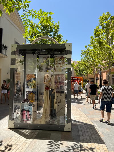 The image depicts an outdoor shopping promenade under a bright, sunny sky, likely in La Roca del Vallès, Spain. In the foreground, a display case shaped like a vintage trunk showcases a mannequin dressed in a flowing cream-colored outfit, sunglasses, and a headscarf, accessorized with a small red purse. The display also includes images of produce, fashion items, and architectural sketches. A woman in a white tank top and white shorts is visible to the left, browsing near the display case.

The promenade features light-colored paving stones and is lined with trees offering dappled shade. Several people are walking along the promenade in the midground and background, some with shopping bags. Buildings with light-colored facades and balconies are visible on either side. The overall atmosphere suggests a vibrant, upscale retail environment.