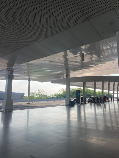 The image depicts an outdoor covered walkway in Ramunia Dua, Indonesia. The walkway is lined with white support columns, and a parking area with motorcycles is visible on the right. A sign with a wheelchair symbol is present. The architecture features a modern design with perforated metal panels on the left. Trees and a bright sky are visible in the background. The floor is reflective, and there are shadows from the columns and other structures.