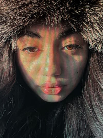 The image is a close-up, low-resolution portrait of a person wearing a fur-lined hood. Sunlight illuminates the face from the left, creating strong shadows and highlights. The individual's eyes are open and looking forward, and their lips are slightly parted. The texture of the fur is visible, and the individual's hair also appears to be long and dark. The background is dark and out of focus. There is no discernible location or context suggesting Chotěšov, Czechia.