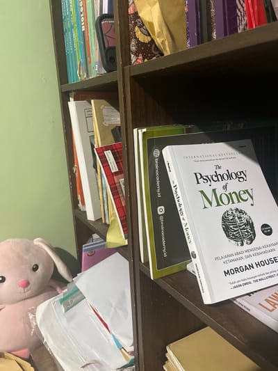 In a dimly lit room, a wooden bookshelf is filled with books and other items. The bookshelf appears to be located in the city of Pasir Gadung, Indonesia. On the top shelf, several books with colorful spines are visible, some with Indonesian titles. A dark-colored object, possibly a phone or camera, is also on this shelf. The middle shelf is more cluttered. A white book with the title "The Psychology of Money" by Morgan Housel is prominently displayed, open to a page with Indonesian text. Next to it are other books, some with plaid covers. In the lower portion of the image, a portion of a pink stuffed bunny is visible. Below the books, on the same shelf, are stacks of papers and folders, some appearing to be schoolwork or documents. A light-colored wall serves as the backdrop on the left side of the image. The overall impression is of a personal space, likely a home or study, with educational materials and personal belongings.