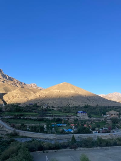 The image depicts a scenic view of the city of Khangkor, India, bathed in the warm glow of what appears to be early morning or late afternoon sunlight. The sky above is a clear, vibrant blue, contrasting with the rugged, golden-brown mountain range that dominates the background. One prominent, conical mountain peak is partially illuminated, casting a long shadow across its slopes.

In the mid-ground, a verdant valley is visible, with patches of cultivated fields and scattered trees. Several buildings are nestled within this landscape. A striking blue-roofed building stands out, flanked by other structures, some with light-colored facades and darker roofs. A cluster of colorful tents, possibly a campground or resort, is situated near a winding road. A small river or stream flows through the valley, crossed by a bridge.

The foreground shows more vegetation, including dense green shrubs and trees, and a paved area with markings, possibly a road or parking lot. The overall impression is one of a tranquil, rural setting within a mountainous region. There are no visible people in the image. The scene is static and peaceful.