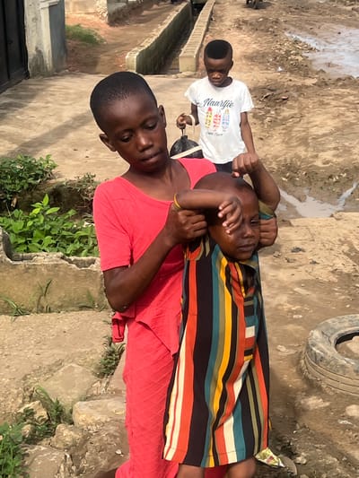 Three individuals are captured in an outdoor setting in Ijegun, Nigeria, during what appears to be daytime with bright, diffused light. In the foreground, an adult, likely female, wearing a red top and a yellow bracelet, holds a younger child. This child is wrapped in a colorful striped garment and has a relaxed expression, with their hands near their face. A silver-colored slipper or sandal is visible on one of the child's feet.

Standing slightly behind and to the right of the adult is another child, wearing a white t-shirt with the word "Naartjie" prominently displayed in cursive, along with smaller, less legible text that includes "Cape Town" and characters like "CH 021." This child also looks towards the viewer and holds a black object, possibly a small bag or strap.

The ground is a mix of dirt and rough concrete or cement, suggesting an unpaved or informal pathway. Scattered patches of green vegetation are visible. In the upper right background, a discarded black rubber tire lies on the ground. Structures that resemble walls or parts of buildings can be seen in the distant background. The scene conveys a casual, everyday moment in a residential area.