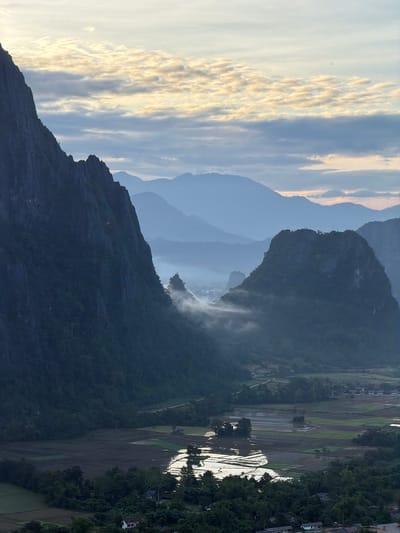 This is a vertical, eye-level, medium shot of a misty, mountainous landscape at dawn. The sky is filled with soft, layered clouds, with streaks of pale yellow and orange suggesting the rising sun. In the foreground, a dramatic, dark, jagged mountain range dominates the left side of the frame, its steep slopes covered in dense green vegetation. To the right, a series of progressively lighter, softer mountain silhouettes recede into the distance, their forms softened by the atmospheric haze. Low-lying mist snakes through the valleys between the mountains, partially obscuring smaller peaks and a distant village. The middle ground reveals a rural scene with patchwork fields, some of which appear to be flooded rice paddies reflecting the sky, and scattered trees. A few buildings are visible nestled among the trees and fields, indicating human habitation. The overall impression is one of serene, natural beauty and a quiet, rural morning. The location is identified as Naka, Laos.