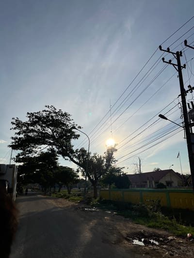 The image depicts a street scene at sunrise or sunset in Alurcucur, Indonesia. The sun is low in the sky, casting long shadows and creating a lens flare in the upper right quadrant of the frame. A large tree with a dense canopy dominates the left side of the image, silhouetted against the bright sky. Power lines crisscross the sky, supported by utility poles. A street lamp is visible on the right side of the road.
On the left side of the street, a vehicle, possibly a truck, is partially visible. Further down the road, a few people on motorcycles can be seen. A yellow and green fence runs along the right side of the street, with a building visible behind it. The road itself appears to be paved but has several potholes and puddles, indicating recent rain or poor road conditions. Vegetation, including grass and some smaller trees, grows along the roadside. A flagpole with a white and red flag, presumably the Indonesian flag, is visible in the distance on the right. The overall impression is of a somewhat rural or suburban street in Indonesia during a transitional time of day.