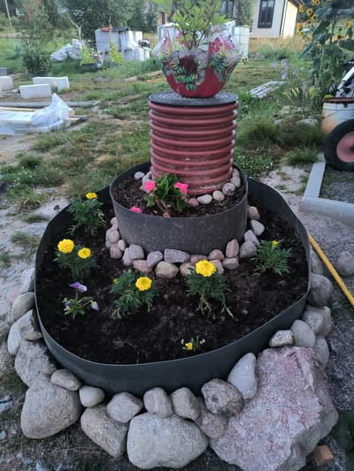 The image shows a raised garden bed constructed from a large red corrugated pipe, possibly a culvert pipe, topped with a black lid. The top of the structure has a planter with a plant and a decorative red pot holding a plant with stones. The main section of the raised bed, which is wider than the top pipe, is filled with dark soil and small yellow and pink flowers. The exterior of the raised bed is surrounded by large and small grey stones. In the background, there are various construction materials, including concrete blocks, and some greenery. The scene appears to be outdoors, possibly in a backyard or a rural setting in Mezhozernoe, Russia, indicated by the construction materials and the naturalistic surroundings. There are no people visible in the image.