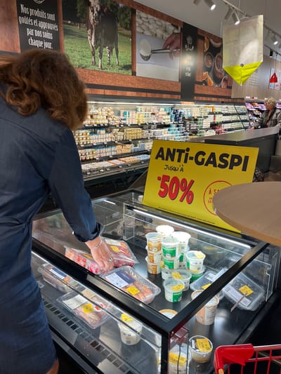 The image depicts the interior of a supermarket in Andrézieux-Bouthéon, France, likely during daylight hours. A woman with reddish-brown curly hair, wearing a dark blue shirt, is seen from the back, reaching into a refrigerated display case. The display case is filled with various food products, including packaged cheeses, yogurts, and possibly seafood. Many of these items have yellow or red discount stickers, indicating a sale.
A prominent yellow sign with bold black text reads "ANTI-GASPI JUSQU'À -50%", advertising a "no waste" initiative with a 50% discount. The sign also mentions "Offre valable sur tous les produits stickés ANTIGASPI, remise déjà effectuée," meaning the offer is valid on all products marked ANTIGASPI, with the discount already applied.
Further in the background, more shelves stocked with dairy products are visible, along with other shoppers browsing. A large mural on the wall features images of cows and agricultural scenery, alongside text in French that translates to "Our products are tasted and approved by us every day." The overall atmosphere suggests a typical grocery shopping experience, with a focus on discounted items. The lighting appears to be artificial, typical of an indoor retail environment.
