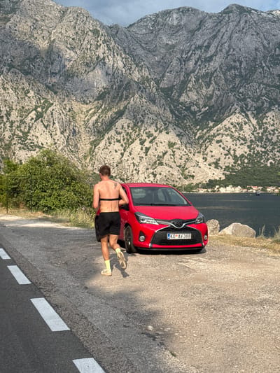A shirtless person with a black heart rate monitor band around their torso is jogging on a paved road with white lane markings. They are wearing black shorts and light-colored socks and running shoes. To the right of the jogger is a red Toyota Yaris with a license plate reading "KO AV 388". The car is parked on a gravel shoulder next to the road. In the background, a dense green bush is visible. Beyond the bush, a large body of dark water, likely a bay or sea, is present, with a distant shoreline dotted with buildings. Towering, rugged, rocky mountains with sparse vegetation form the backdrop. The sky is partly cloudy. The scene appears to be in Dražin Vrt, Montenegro, given the mountainous coastal landscape.