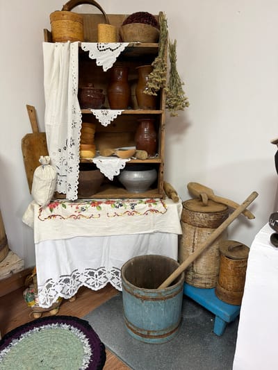 This is a forensic, matter-of-fact description of a staged domestic interior scene, likely in Ust'-Pyzas, Russia, depicting a collection of rustic, traditional household items. The scene is lit by ambient light, creating shadows that add depth to the objects.
The central focus is a wooden shelving unit filled with various earthenware and wooden vessels. On the top shelf, a large wooden bowl contains dark red material, possibly dried berries or grain. Next to it are two birch bark containers, one larger than the other. A wooden-rimmed basket is positioned to the left.
The shelves below are adorned with white lace-trimmed cloths. Earthenware jugs and pots of various sizes and shapes are displayed prominently, some with a glossy finish, others matte. There are also stacks of small wooden bowls and cups, and a larger metal pot on one of the lower shelves. A rolled-up white cloth hangs on the left side of the shelving unit, partially obscuring the view of some items.
To the left of the shelving unit, a wooden paddle or board leans against the wall. In front of the shelving unit, a small white cloth bag is tied at the top. Below the shelving unit, a white embroidered tablecloth with floral and berry motifs is draped.
On the floor in front of the shelving unit and to the right, several other items are arranged. A light blue wooden bucket with metal banding contains a long wooden pole. Behind this bucket are two larger wooden cylindrical containers, possibly butter churns or storage containers, with one having a wooden lid and a large wooden spoon resting on it. A small blue wooden stool supports one of these cylindrical containers. To the far right, a white surface, possibly a table or counter, is visible, with some dark objects on it. A circular rag rug in shades of green, purple, and black is situated on the floor to the lower left.
The overall impression is one of a carefully curated display of historical or traditional household artifacts, evoking a sense of rural life. There are no people present in the scene, and no actions or interactions are depicted. The environment appears to be an indoor setting with a plain white wall in the background and wooden flooring.