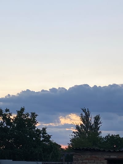 A daytime shot captures a sky with soft blue and white hues. The horizon is marked by a band of dark, puffy clouds, with patches of orange light visible through gaps. In the foreground, the dark green foliage of trees is prominent, partially obscuring a brick structure with a corrugated roof. The overall impression is of an outdoor scene in Rozkvit, Ukraine, likely during the late afternoon or early evening. No people are visible.