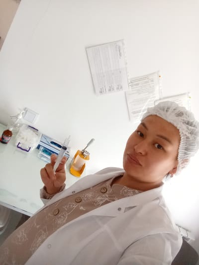 A selfie is taken from a low angle in a room that appears to be a medical or laboratory setting. The subject is a woman wearing a white disposable hairnet and a white lab coat over a tan garment with a floral pattern. She is looking directly at the camera with a neutral expression. In her right hand, she is holding a syringe. On the table in front of her, there are various medical supplies, including a bottle of liquid, cotton balls in a container, a box of gloves, and a sharps container. The wall behind her has several documents pinned to it. The lighting in the room is bright, suggesting it may be daytime or have strong artificial lighting. The environment suggests a professional medical or scientific setting, potentially in a clinic or research facility. The specific location in the city of Жошук, Kyrgyzstan, cannot be determined from the visual information provided in the image.