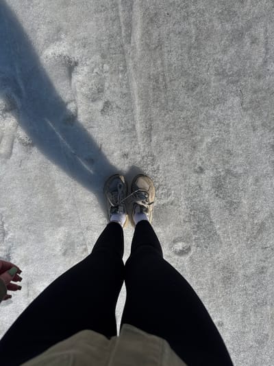 A forensic, top-down view depicts an individual standing on a surface of granular ice or compacted snow in Oleshkovo, Russia. The subject wears black, ribbed leggings, and light grey athletic shoes over white socks. A left hand with green painted fingernails is partially visible in the lower left quadrant. A distinct dark shadow extends diagonally from the upper left, falling across the reflective, frozen ground, indicating direct sunlight.
