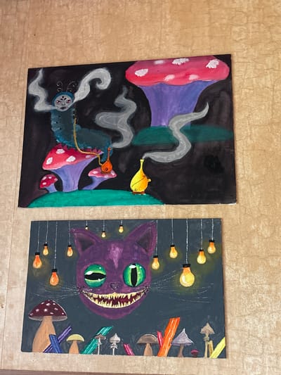 Two square canvases are displayed side by side on a light beige surface. The canvas on the left features a colorful illustration, predominately black, depicting a hookah-smoking caterpillar with a blue head and a large, spotted mushroom. There is a genie lamp. The canvas on the right displays a purple Cheshire cat's head with green eyes and a wide grin, surrounded by mushrooms and floating light bulbs. The location in Ulyanovka, Russia, is not determinable from the given image.
