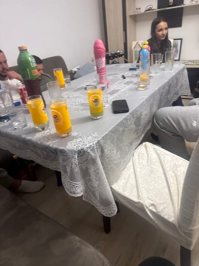 The image shows a group of people seated around a table covered with a white lace tablecloth. The table is cluttered with glasses and bottles of various beverages, including orange juice, soda, and mineral water. A black mobile phone sits on the table. People are seated around the table, and one is wearing grey sweatpants with a New Balance logo. The location could be Graf Ignatievo, Bulgaria, but there is no evidence in the image to confirm this.