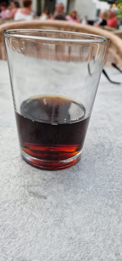 A close-up, vertical, eye-level shot focuses on a partially filled glass of dark liquid, likely soda or iced tea, resting on a textured, light-grey surface, possibly a table. The glass is clear and shows some condensation on the outside. The liquid is a deep amber color, with lighter hues where the light refracts through it, creating a warm glow at the bottom. The background is softly blurred, revealing indistinct figures of people seated at tables, suggesting an outdoor dining or social setting. The overall atmosphere is casual and relaxed. The specific location in Las Rosas, Spain, is not determinable from the image alone.