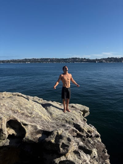 A lone male stands shirtless in black swim shorts on a weathered, light-colored rock outcrop overlooking the deep blue water of Rose Bay, Australia. He is barefoot, facing the water, with his head tilted upwards towards a clear blue sky and arms outstretched, palms open. The individual possesses a muscular physique and wears a gold-colored pendant necklace. The rock formation in the foreground exhibits natural erosion and sun-bleached patches. In the mid-distance, a white sailboat navigates the bay. The distant shoreline reveals a cityscape of buildings and dense foliage, interspersed with numerous moored boats and yachts. The scene is brightly lit by direct sunlight.