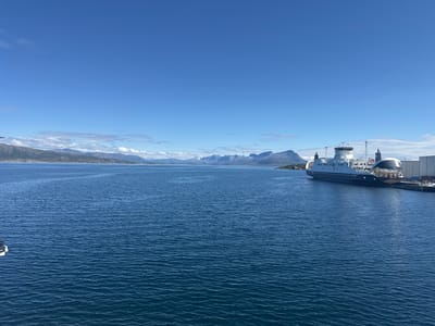 This media file captures a serene view of a ferry docked at a harbor, likely in Lødingen, Norway. The clear blue sky and calm, deep blue water suggest a pleasant day, possibly midday due to the bright sunlight. In the background, majestic mountains rise, some with snow-capped peaks, hinting at a northern, possibly Arctic or sub-Arctic, environment. The ferry itself is a prominent object, a large vessel with multiple decks and a bridge structure. It appears to be stationary at a pier, indicated by the docking facilities and a building in the background. The water has a gentle ripple, reflecting the clear sky. The overall mood is one of tranquility and natural beauty, with the ferry being a man-made element integrated into a stunning natural landscape. There is no visible text in the image.
