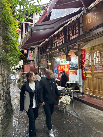 This image presents a narrow, wet stone alleyway in Gongtan, China. In the immediate foreground, a woman with light brown hair, dressed in a white t-shirt and a two-tone black and white puffer jacket, walks hand-in-hand with a bald man wearing a black puffer jacket featuring a white 'NY' logo, black pants, and grey/white sneakers. Both are moving away from the camera, with the woman smiling and looking to her right, and the man looking straight ahead.

To the left, a rough-hewn stone wall is densely covered with green ferns and other foliage. A red lantern is partially visible hanging from the wall further back. To the right, traditional multi-story wooden buildings feature intricate lattice window screens and support maroon retractable awnings. Numerous signs with Chinese characters are affixed to these buildings, including a large carved wooden sign overhead that reads "田家" and brightly lit signs advertising "鲜肉饼" (fresh meat pies) along with phone numbers. Several red lanterns and hanging baskets with plants are suspended from the wooden structures. Further down the alley on the right, a woman in a dark uniform stands behind an outdoor food preparation counter equipped with metal bowls, a table, and chairs. The ground is paved with uneven flagstones, which appear wet and reflective. The ambient lighting is diffused, suggesting an overcast day or evening.