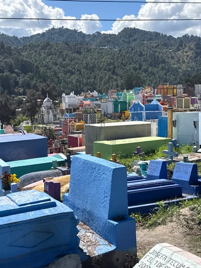 This vibrant, colorful cemetery, likely in Guatemala or Mexico, stretches up a hillside near a dense forest. Tombstones are painted in blues, greens, yellows, and reds. The names Marta Panjot Perez and Josefa Tecums are visible on some stones, with death dates in 1983 and 2003, respectively. The scene is captured under a bright sky with fluffy clouds.