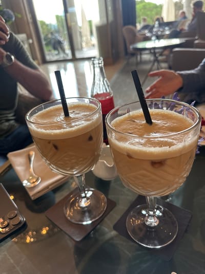 The image captures a detailed view within a high-end lounge or cafe in Lausanne, Switzerland. In the foreground, two stemmed glasses containing iced coffee with a frothy top and visible ice cubes are positioned centrally on a reflective dark table. Each glass holds a black drinking straw and rests on a dark, square coaster.

To the left of the glasses, a silver-colored smartphone is partially visible on the table. Adjacent to it, a light-colored, possibly leather, folio or folded napkin holds a silver spoon. A clear glass Coca-Cola bottle with a red label, appearing partially consumed, stands behind the two coffee glasses. A small white ceramic creamer is situated near the base of the right coffee glass.

In the mid-ground, partial views of two individuals are present. On the left, an arm wearing a green shirt and a metallic wristwatch is visible. On the right, a hand is extended, possibly mid-gesture.

The background reveals a spacious interior with large glass doors or windows, through which an outdoor scene with blurred greenery and other patrons seated at tables is visible. The indoor environment features additional tables and upholstered chairs, consistent with a luxury lounge. The floor appears to be a light, reflective material.