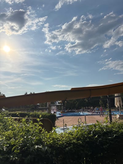 The image depicts an outdoor swimming pool facility on a sunny day with scattered clouds. In the foreground, lush green bushes obscure the lower portion of the scene. A large, yellow water slide extends horizontally across the middle of the frame, casting a shadow. Beyond the slide, several people are visible, some interacting with a pool area where water is being sprayed, suggesting water features or a slide exit. Others are seen near the edges of different pools, and one person is walking along the poolside. The background features trees and other elements of the park, under a bright blue sky. The scene is likely located in Bürgstadt, Germany, given the context provided, though specific landmarks to confirm this are not visible in the image.