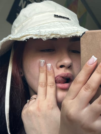 The image shows a close-up, vertically oriented shot of a young person's face and hands. The person is wearing a cream-colored bucket hat with frayed edges and a small metal embellishment on the side. Their fingernails are painted with a French manicure. The person's mouth is open, with their tongue slightly visible. One hand is positioned near their mouth, obscuring part of their face, and the other hand is resting near their chin, showing a ring with a small stone on one finger. The background is blurred, but hints of dark hair and possibly clothing with some visual texture are visible. The lighting suggests an indoor setting. The overall mood is playful and perhaps suggestive due to the pose. No specific time of day or weather cues are apparent. The location context of Starobaltachevo, Russia, is provided externally and is not visually discernible from the image itself.