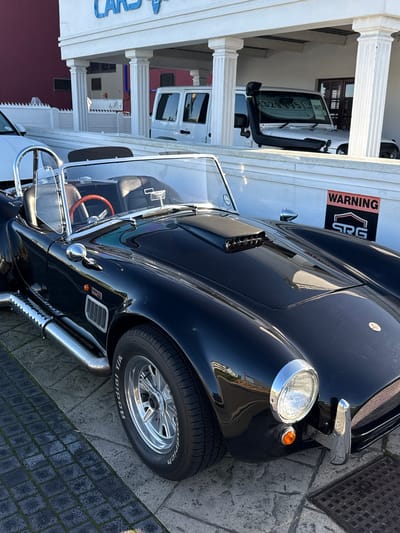 The image shows a black AC Cobra car parked on a paved area in front of a building that appears to be a car dealership. The building has white columns and a sign that partially reads "CARS 4". To the right of the Cobra, two white Jeep Wranglers are visible parked under the building's overhang. One of the Jeeps has a snorkel attached to its exterior and a price tag of "R329 995" displayed on its windshield. A red "WARNING" sign with the logo "SRG" is affixed to the wall behind the Cobra. The car dealership is located in Blouberg, South Africa, as indicated by the presence of a South African flag logo on one of the vehicles. The lighting suggests it is daytime, with sunlight casting reflections on the vehicles.