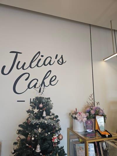The image depicts a decorated Christmas tree in front of a white wall with black lettering that reads "Julia's Cafe." The Christmas tree is adorned with ornaments, including bells, baubles, and a star topper. To the right of the tree, on a glass-topped table, are two awards, a vase of flowers, and several books on a lower shelf. The awards appear to be commemorative or recognition plaques, one of which has a purple base. The flowers are a mix of light pink and lavender hues. The books are various titles, some with visible spines. The overall setting suggests a cozy and festive atmosphere, possibly within a cafe or shop. The specific location is identified as Banyuurip, Indonesia, suggesting this is an establishment within that city. There are no people visible in the image, nor any explicit actions or interactions taking place. The environment is well-lit, with artificial lighting visible from a fixture on the ceiling.